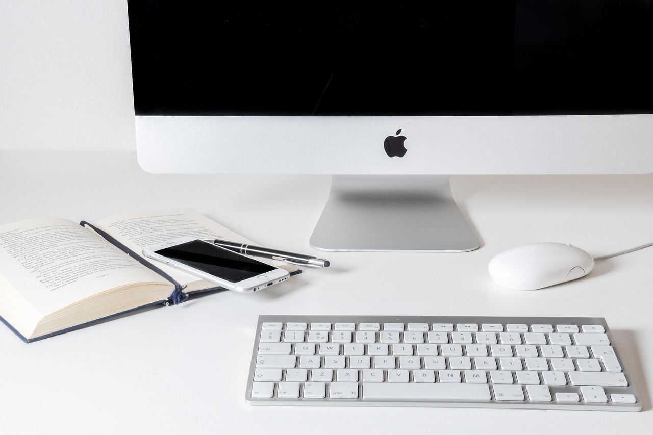 A desk with a monitor, a keyboard, an open book, a phone and a pen