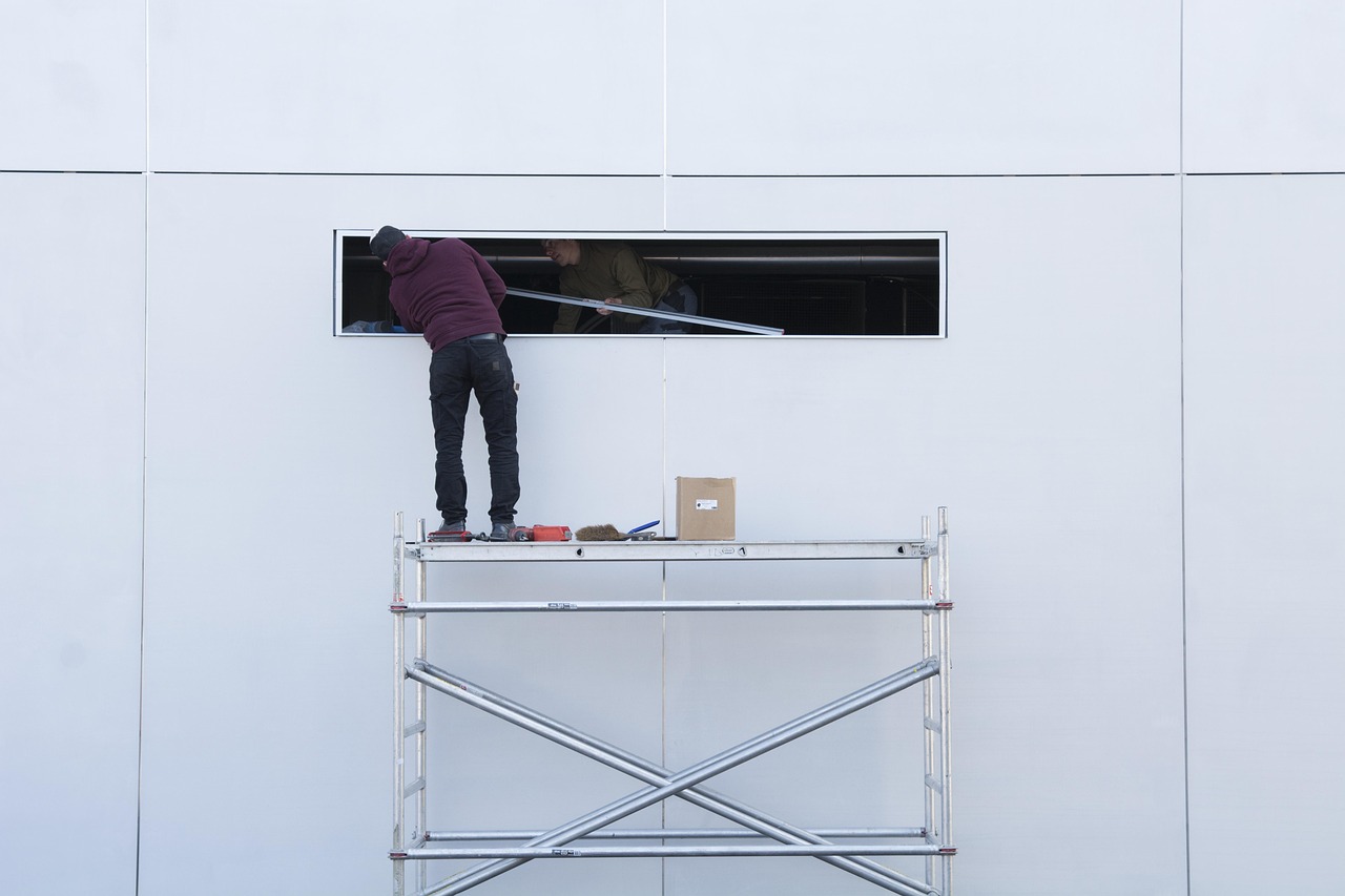 Two workers on scaffolding fitting a long trim board into a window opening on a white panel wall